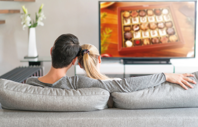 Pärchen auf der Couch sieht Pralinenbote im Fernsehen